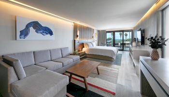 A modern hotel suite with a sofa and coffee table, a neatly made bed, and ocean-view windows under warm ambient lighting.