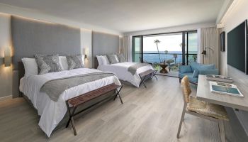 A bright modern hotel bedroom with twin beds, pillows, and a balcony view over the ocean and palm trees.