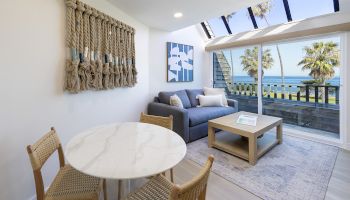A bright living room with a blue sofa, round marble table, woven chairs, and a large ocean-view window, styled with coastal decor.