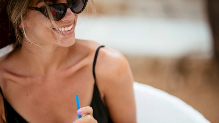 A woman in sunglasses smiles while holding a bright blue straw on a bed, as if sipping a drink in a relaxed, sunny vibe.