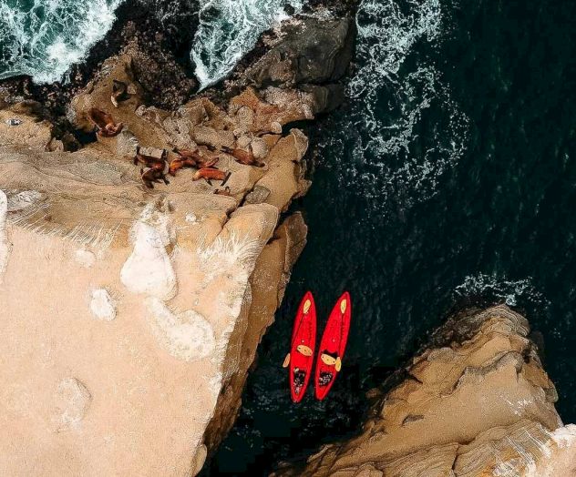 Cliffside shore with two red kayaks resting on a rock ledge above dark blue water crashing below. A daring cliffside view.
