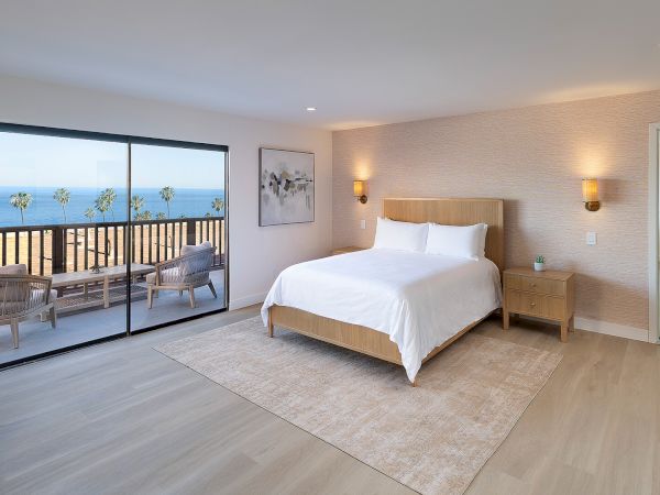 Modern hotel room with a large bed, soft beige tones, and a glass sliding door to a balcony overlooking the ocean; light wood floors and a view into a bright adjoining room.