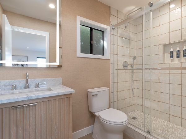 A modern bathroom with a double-sink vanity, wide mirror, beige walls, and a glass-enclosed tile shower next to a toilet.