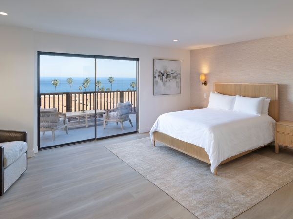 A bright hotel room with a large bed, nightstands, a gray sofa, and a balcony overlooking water; airy, modern decor and soft lighting.