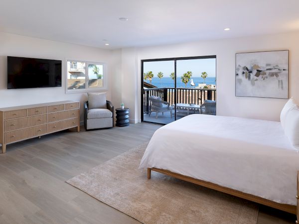 A bright hotel-style bedroom with a large bed, wood furniture, a TV console, a seating area, and a balcony view of palm trees, all in soft neutrals.