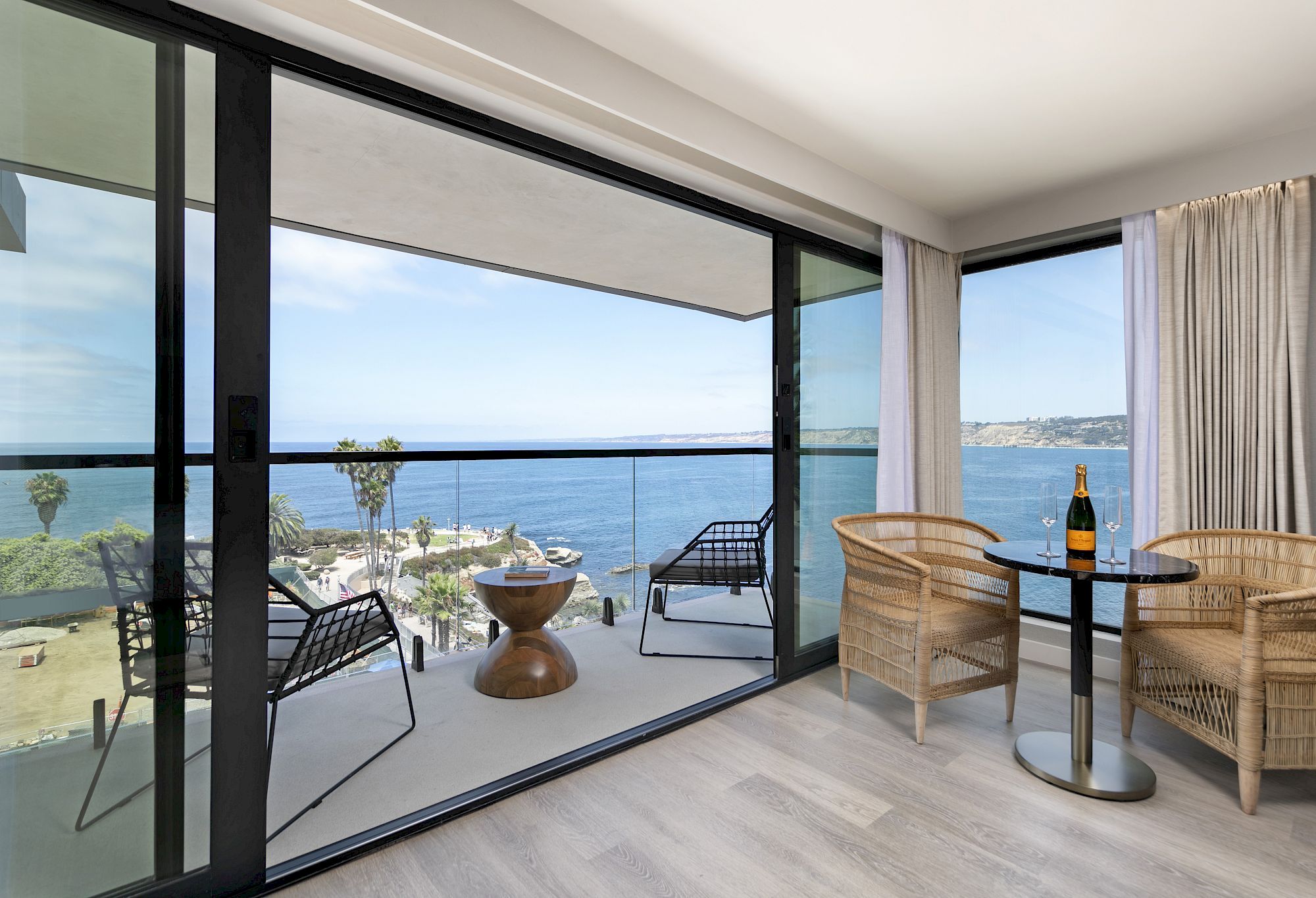 A modern seaside balcony with woven chairs and table, watching the ocean through floor-to-ceiling windows.